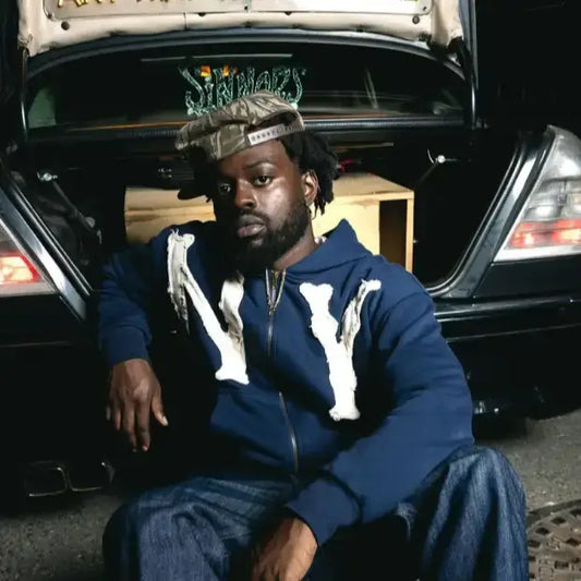 Man in blue outfit and crown by car trunk showcasing 2000s hoodie with embroidered design