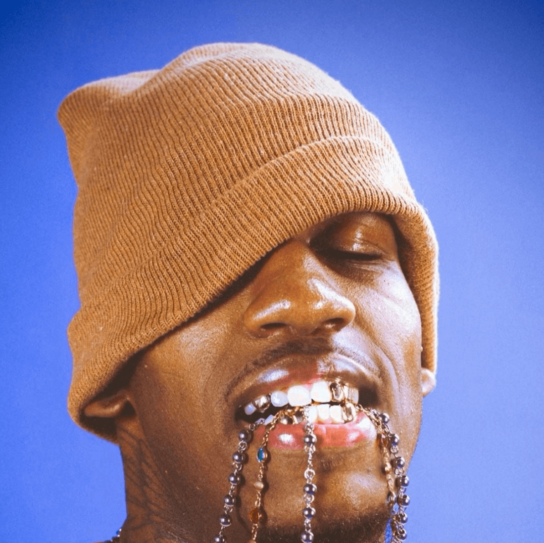 Person wearing a tan beanie pulled over their eyes, with decorative beads in their mouth, set against a blue background