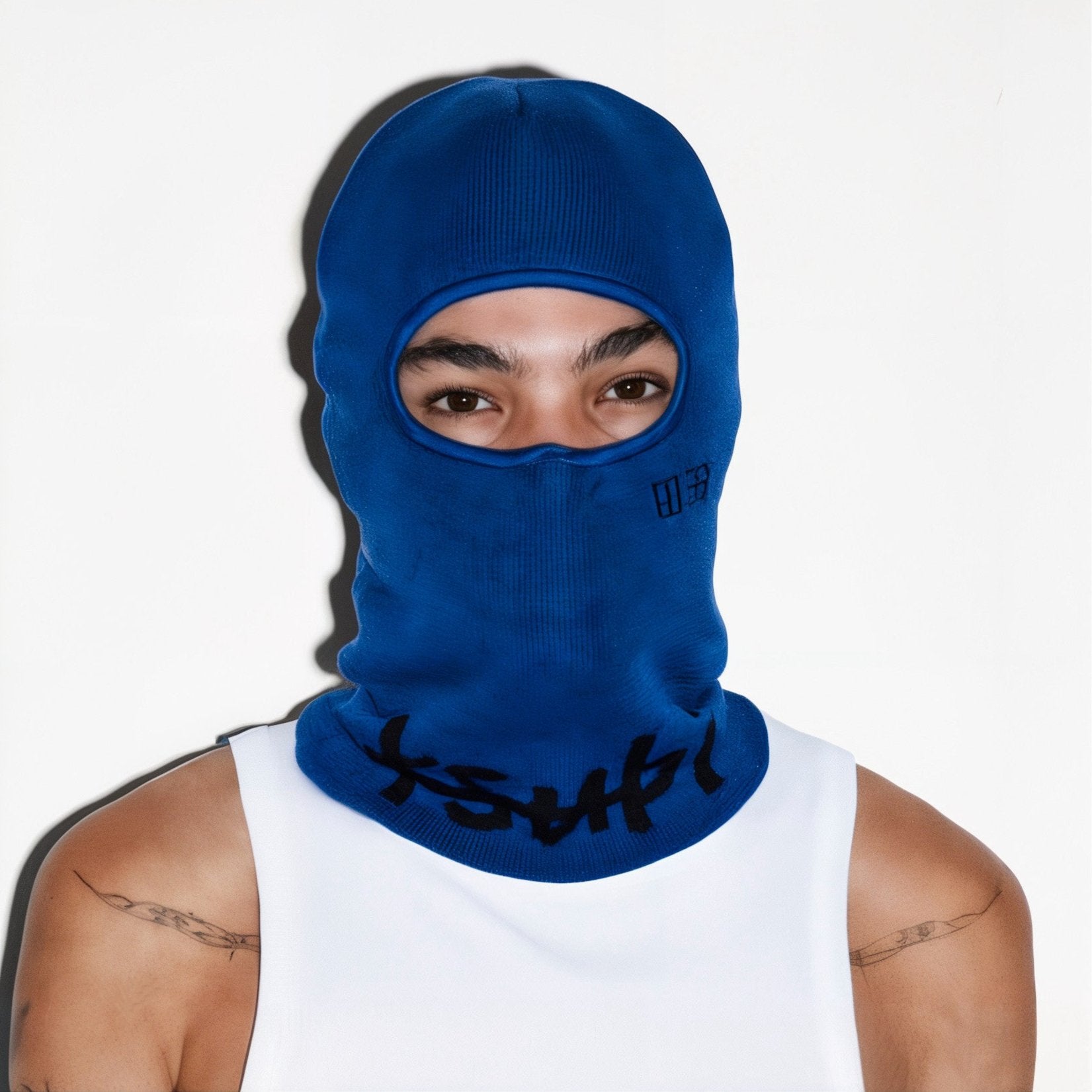 Person wearing a blue balaclava with an open eye area, dressed in a white tank top against a plain background