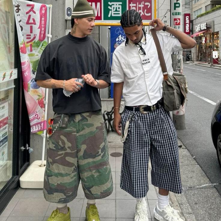 Two men in Y2K streetwear: camo shorts, black tee, and checkered shorts, white shirt