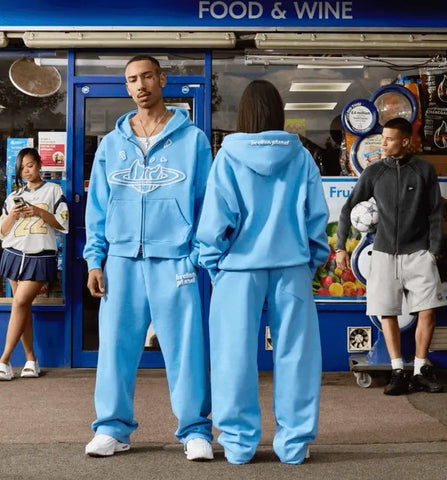 Person in light blue Nike tracksuit demonstrating oversized hoodie fit in front of store.