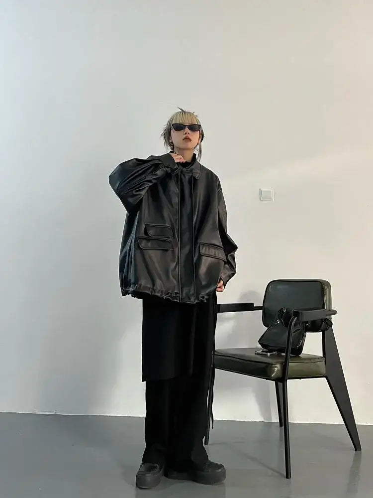 Person in oversized leather jacket and sunglasses next to a chair in stylish outfit